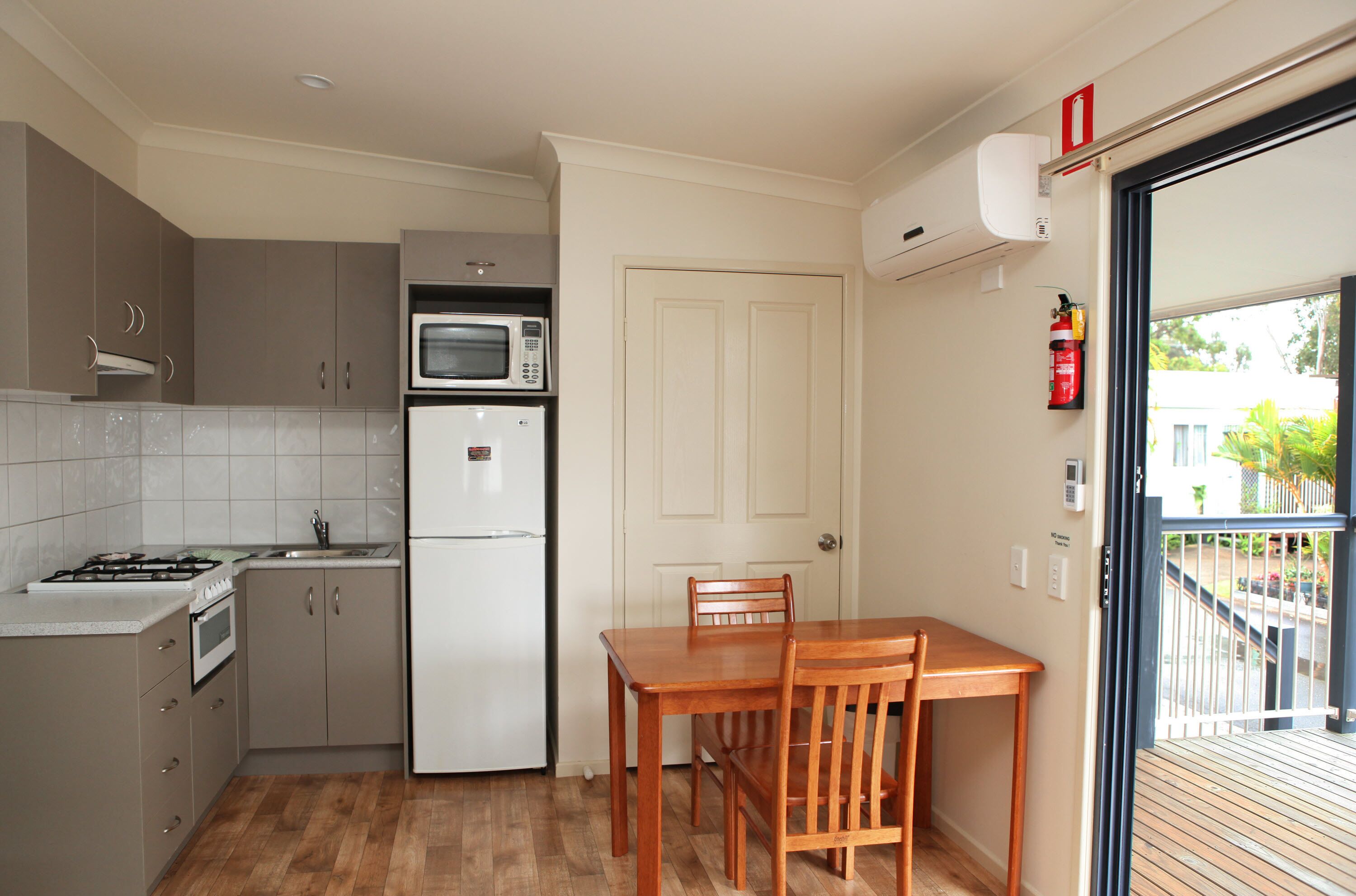 A compact kitchen with a gas stove, microwave, and refrigerator, a wooden dining table with chairs, and a door leading to a balcony with a view of greenery.