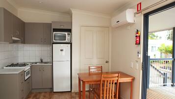 A compact kitchen with a gas stove, microwave, and refrigerator, a wooden dining table with chairs, and a door leading to a balcony with a view of greenery.