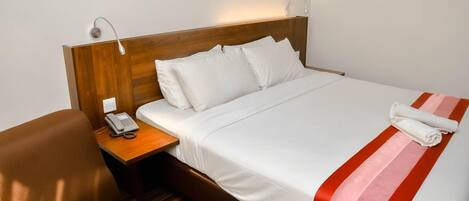 A hotel room with a wooden headboard, a bed with white bedding and a red and pink striped blanket, a brown armchair, a wooden nightstand with a telephone, and a wall-mounted lamp.