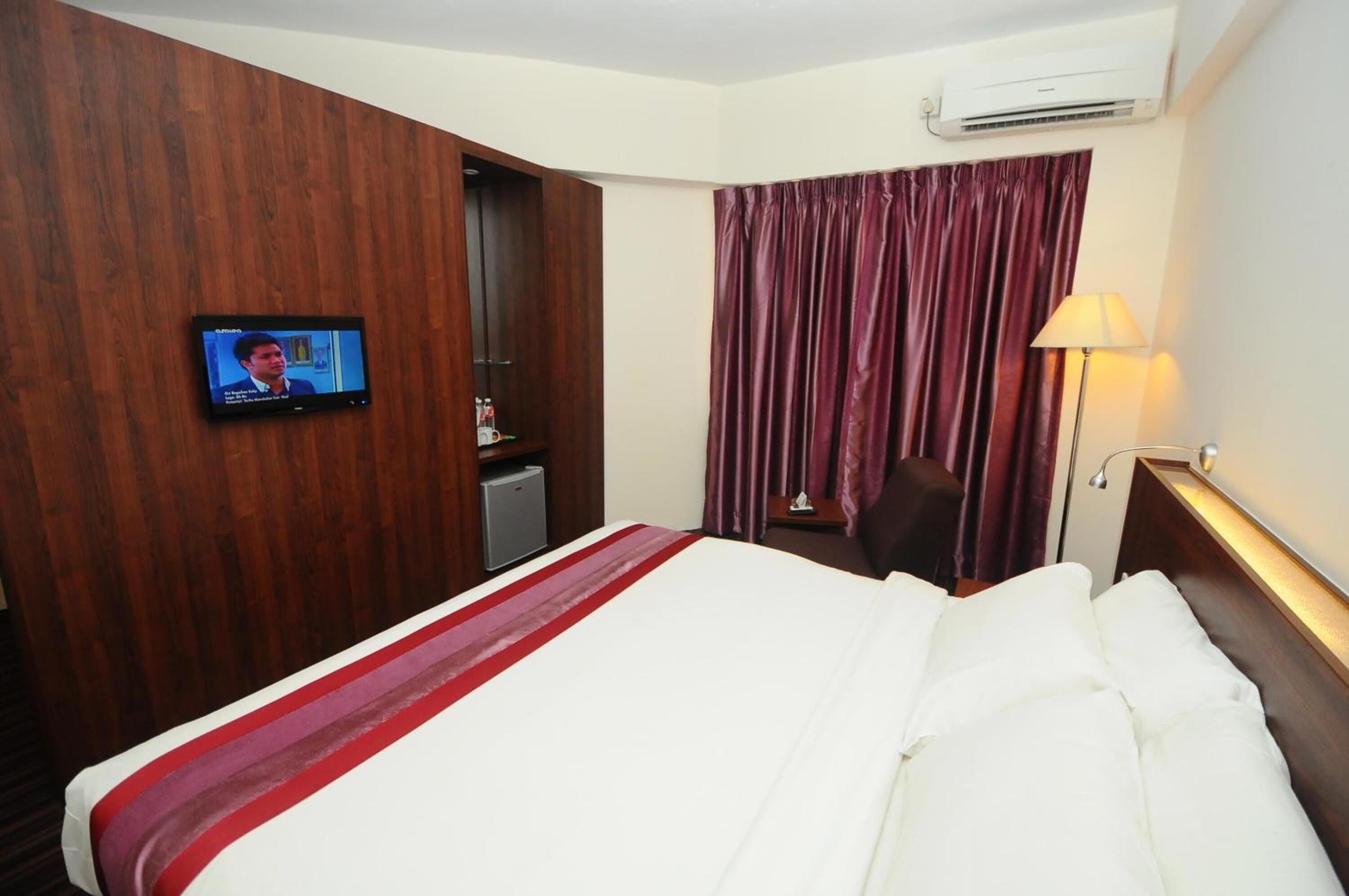 A hotel room with a bed, a television on the wall, a desk with a chair, and a window with curtains.