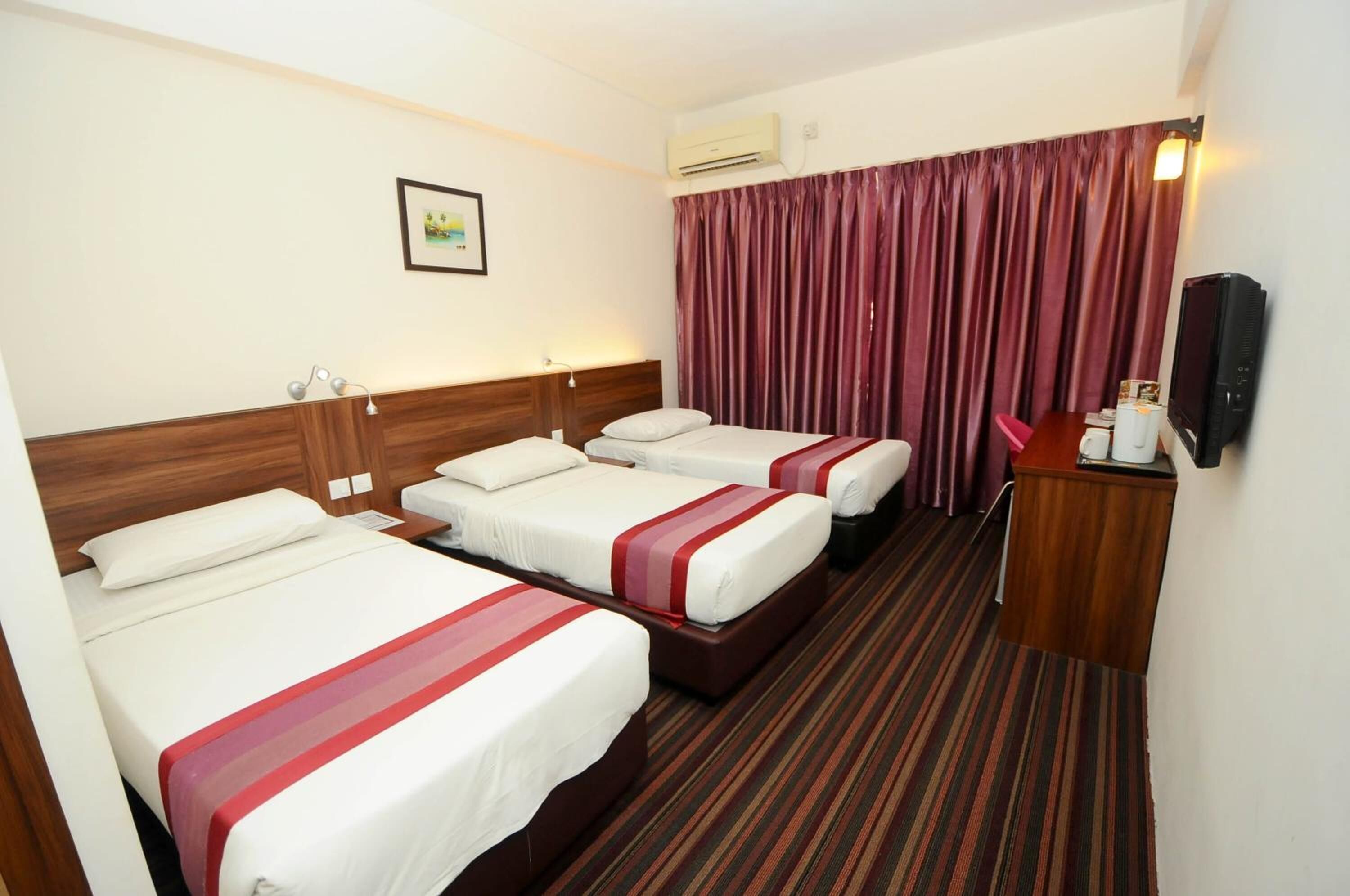 A hotel room with three beds, a wooden headboard, a flat-screen TV, a small table with a coffee maker, and a window with curtains.