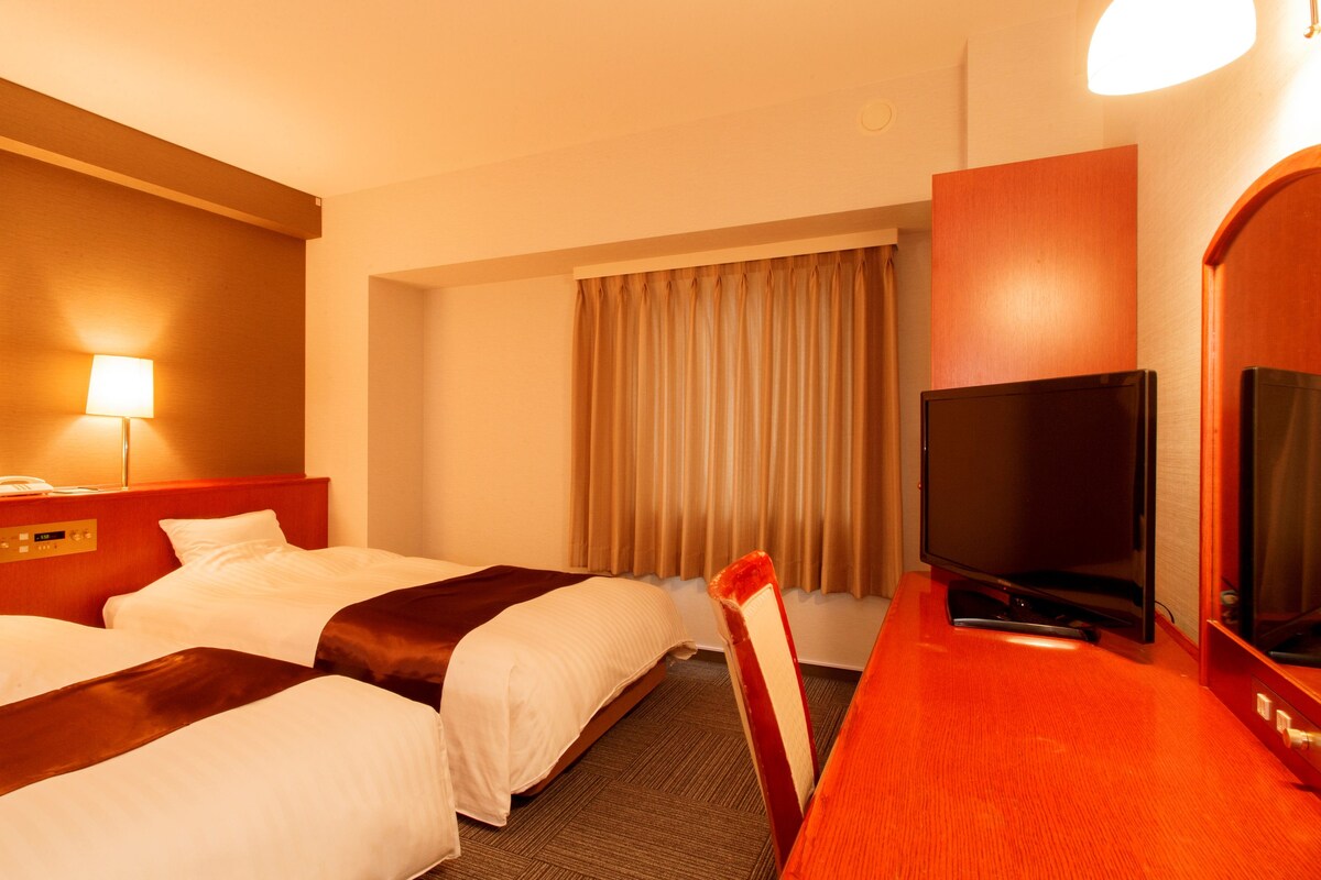 A hotel room with two beds, a desk with a TV, and a window with curtains.