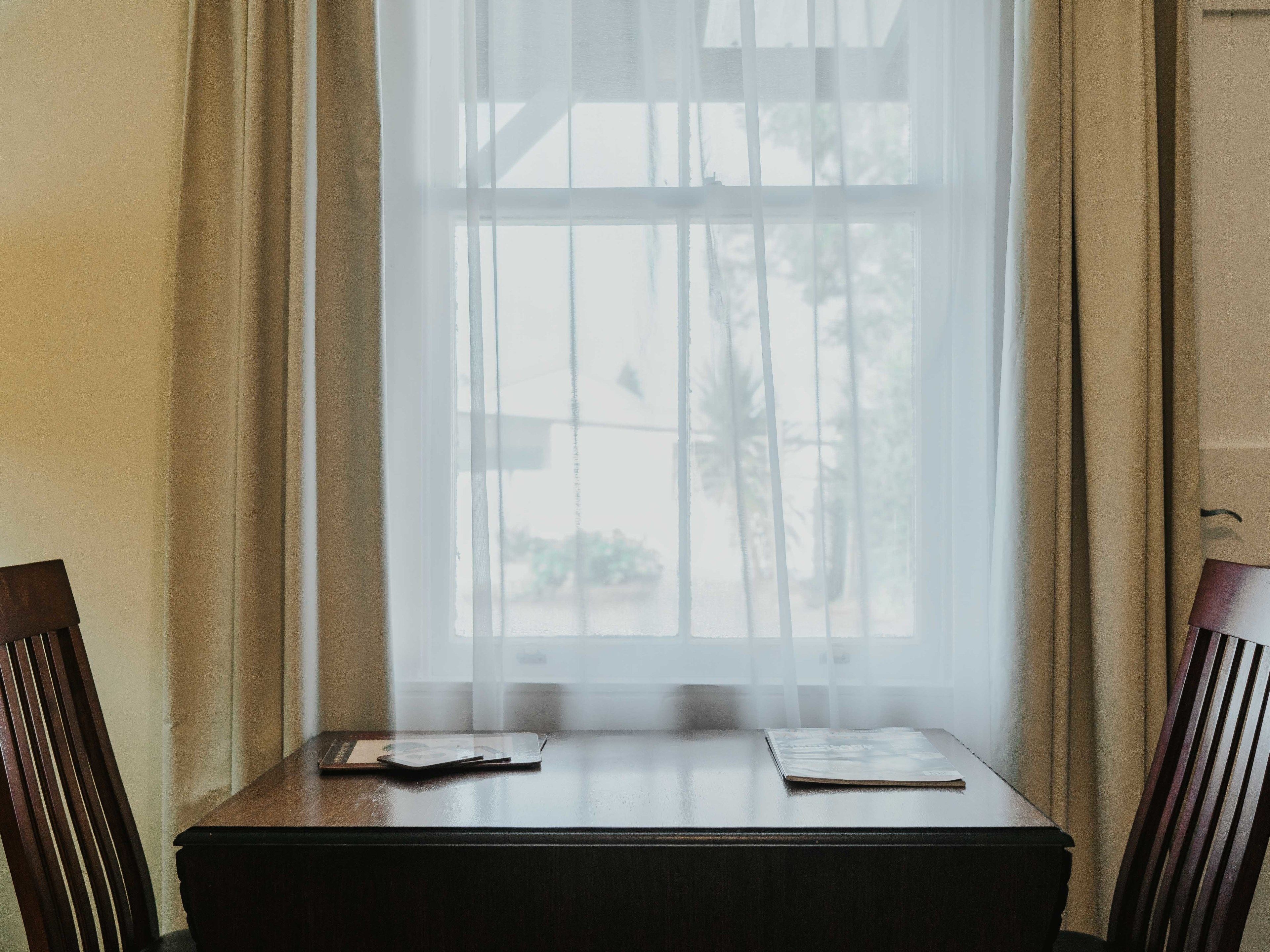 Une pièce avec une table et deux chaises, une fenêtre avec des rideaux et une vue sur des palmiers à l’extérieur.
