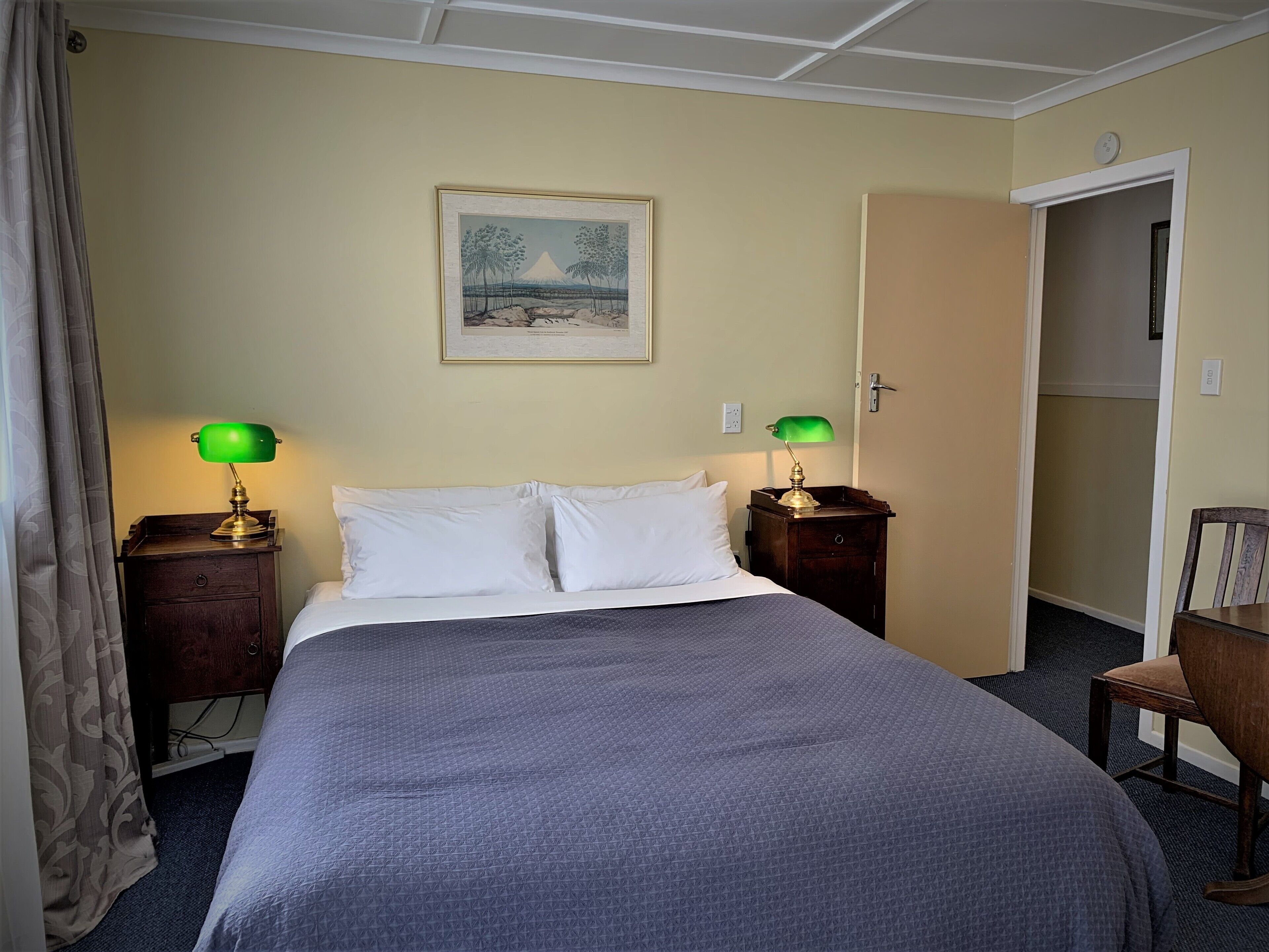 Habitación de hotel con una cama, dos mesitas de noche con lámparas, un cuadro en la pared y una puerta a otra habitación.