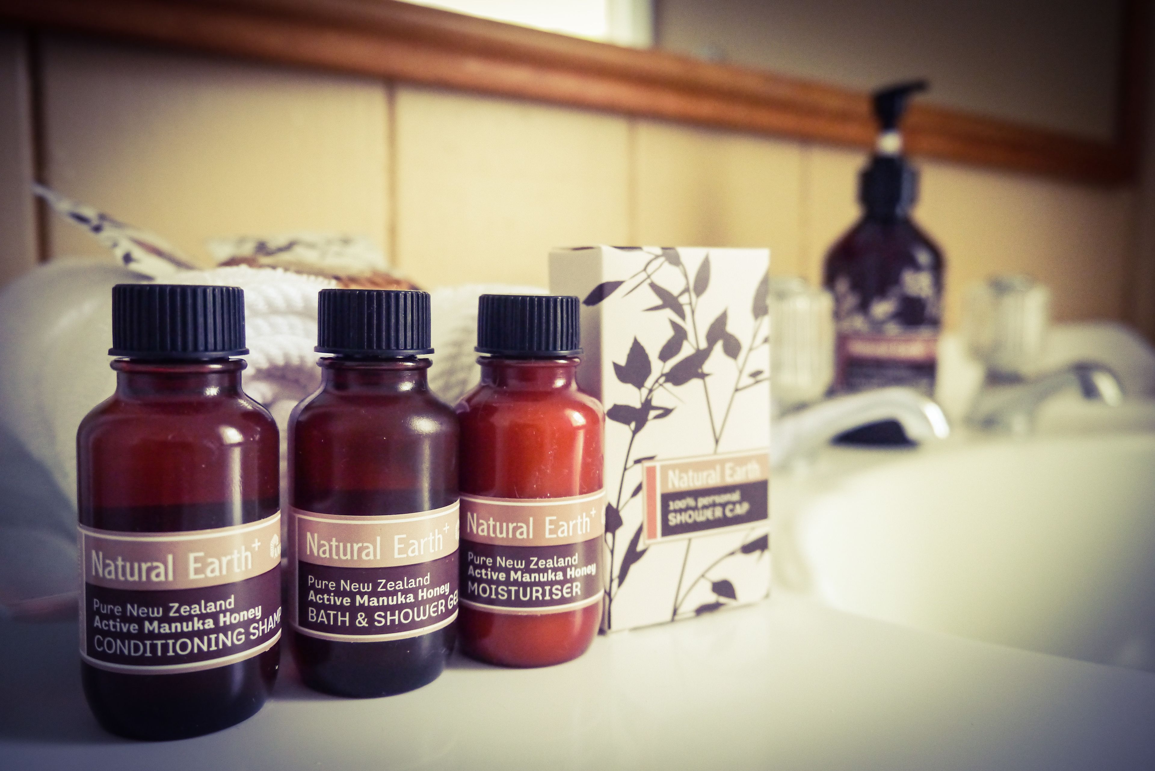 Three bottles of Natural Earth+ bath and shower products and a shower cap box on a bathroom counter.