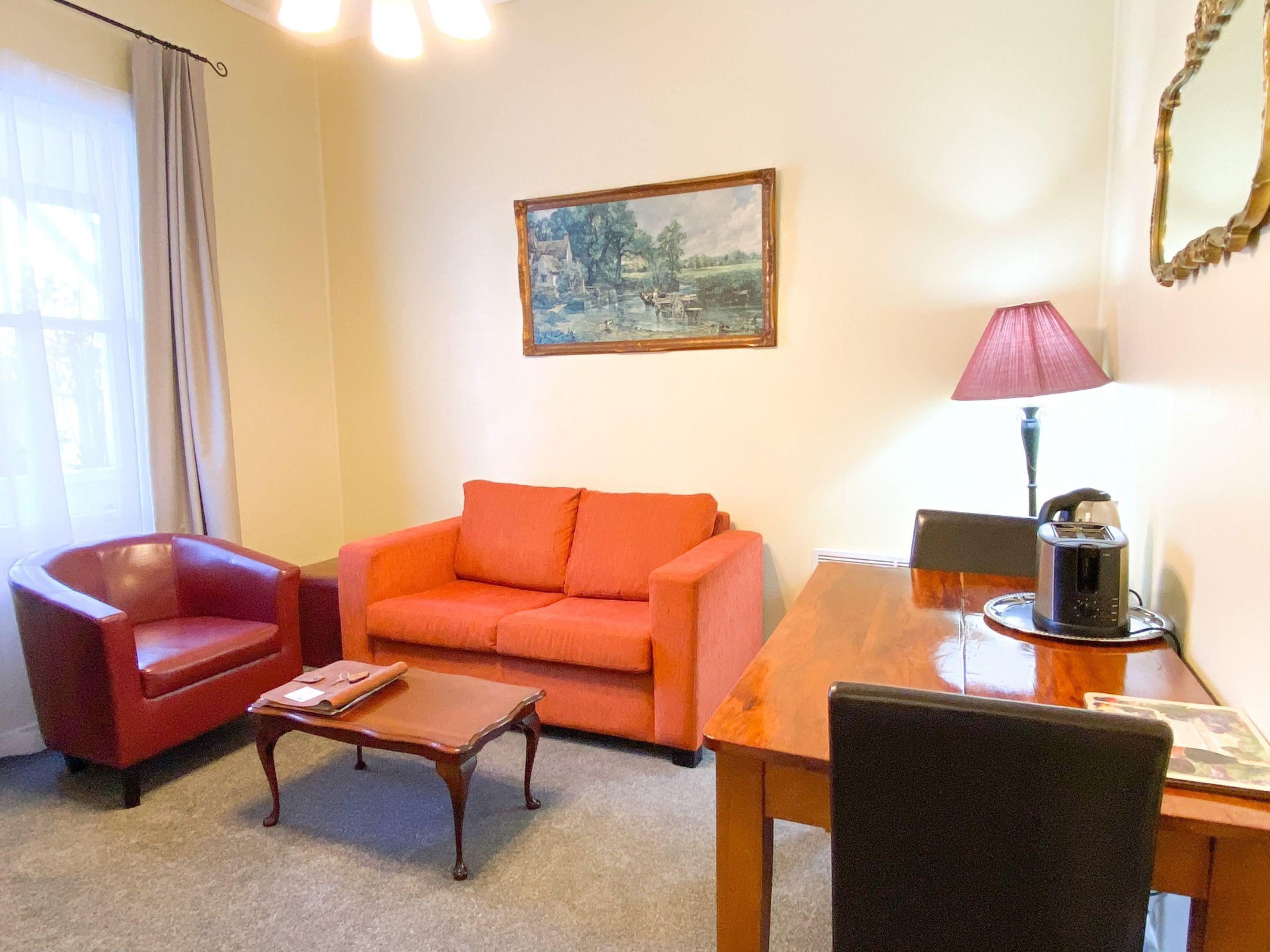 A living room with a red sofa, a wooden coffee table, a red armchair, a wooden dining table, a lamp, and a painting on the wall.