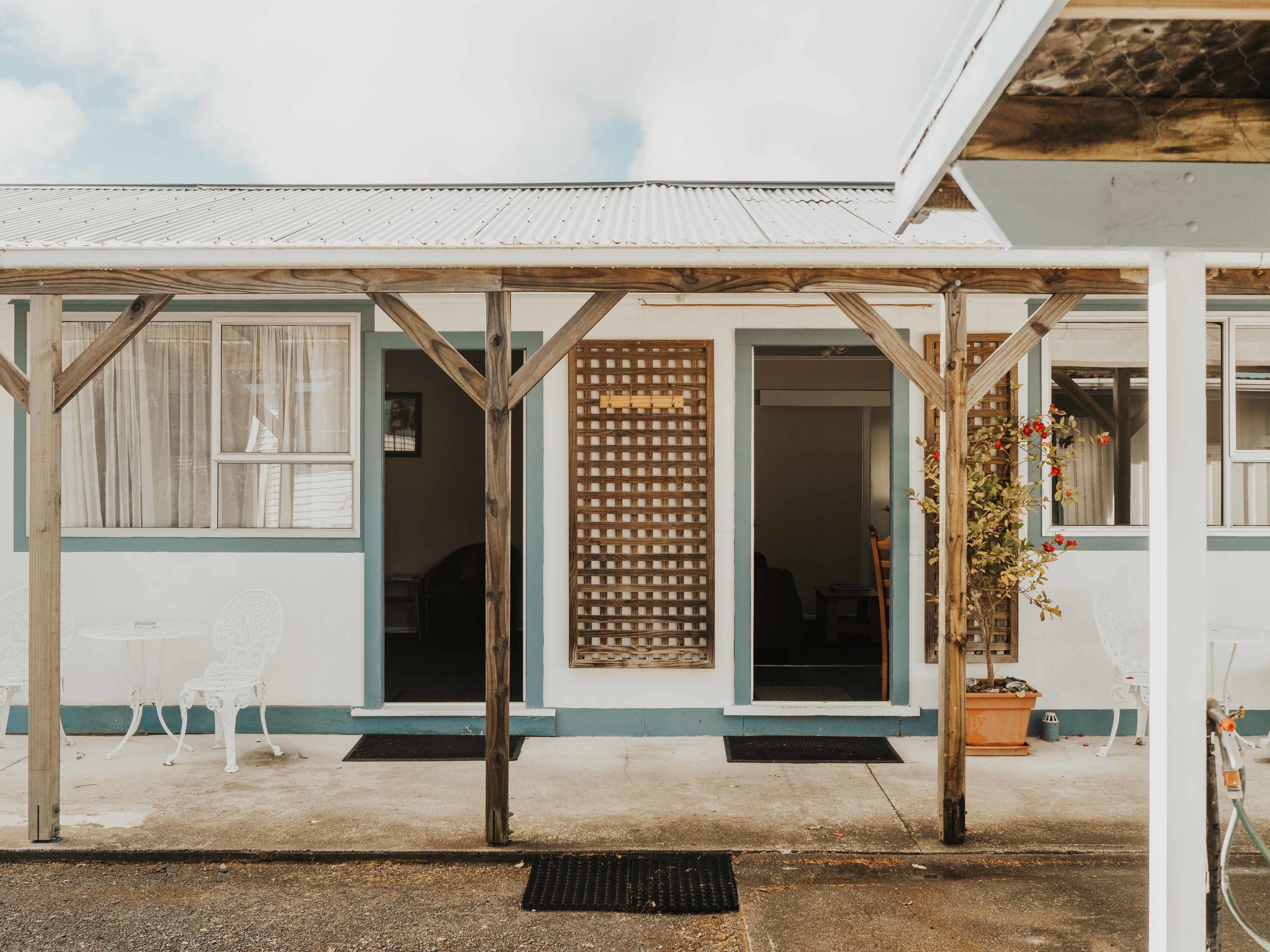 A building with a wooden porch supported by beams, a corrugated metal roof, and a door with a wooden lattice screen.