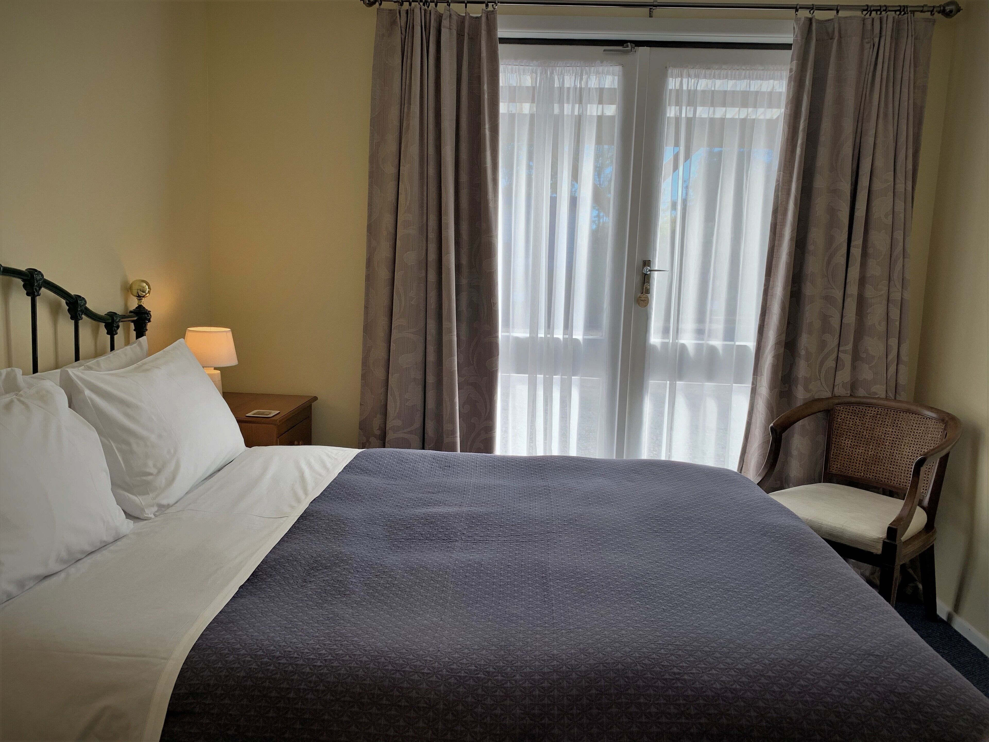 A neatly made bed with white and grey linens, a wooden nightstand with a lamp, a chair, and a window with curtains.