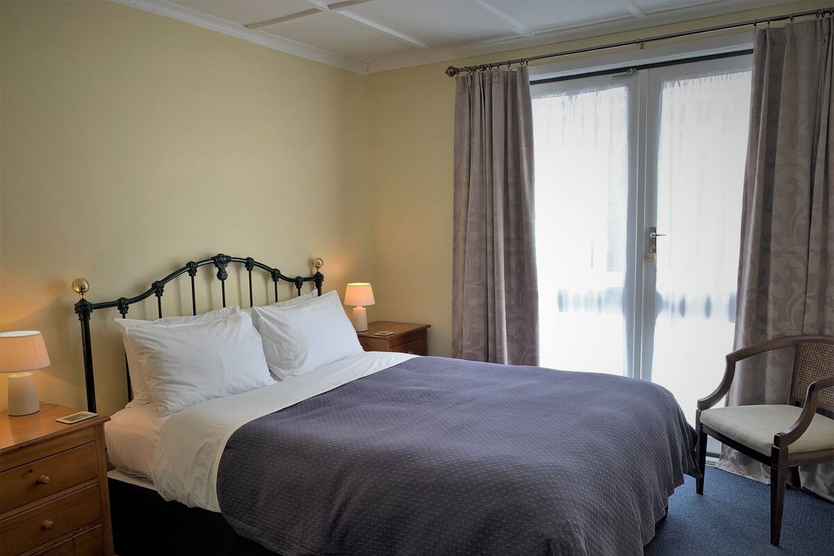 Een net opgemaakt bed met witte en grijze lakens, een houten nachtkastje met een lamp, een stoel en een raam met gordijnen.