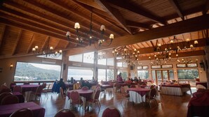 A spacious indoor dining area with high wooden ceilings and multiple chandeliers. Tables are arranged for groups, and large windows provide views of the outdoors. - Amancay (San Carlos de Bariloche)