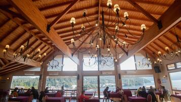Un restaurante con vigas de madera y candelabros, ventanas amplias con vistas a las montañas y mesas dispuestas para comer.