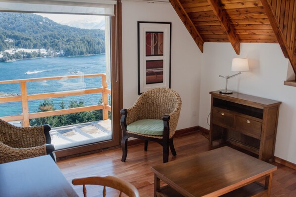A room with a wooden ceiling, a view of the sea, a wicker chair, a wooden table, and a desk with a lamp. - Intersur Amancay Bariloche (San Carlos de Bariloche)