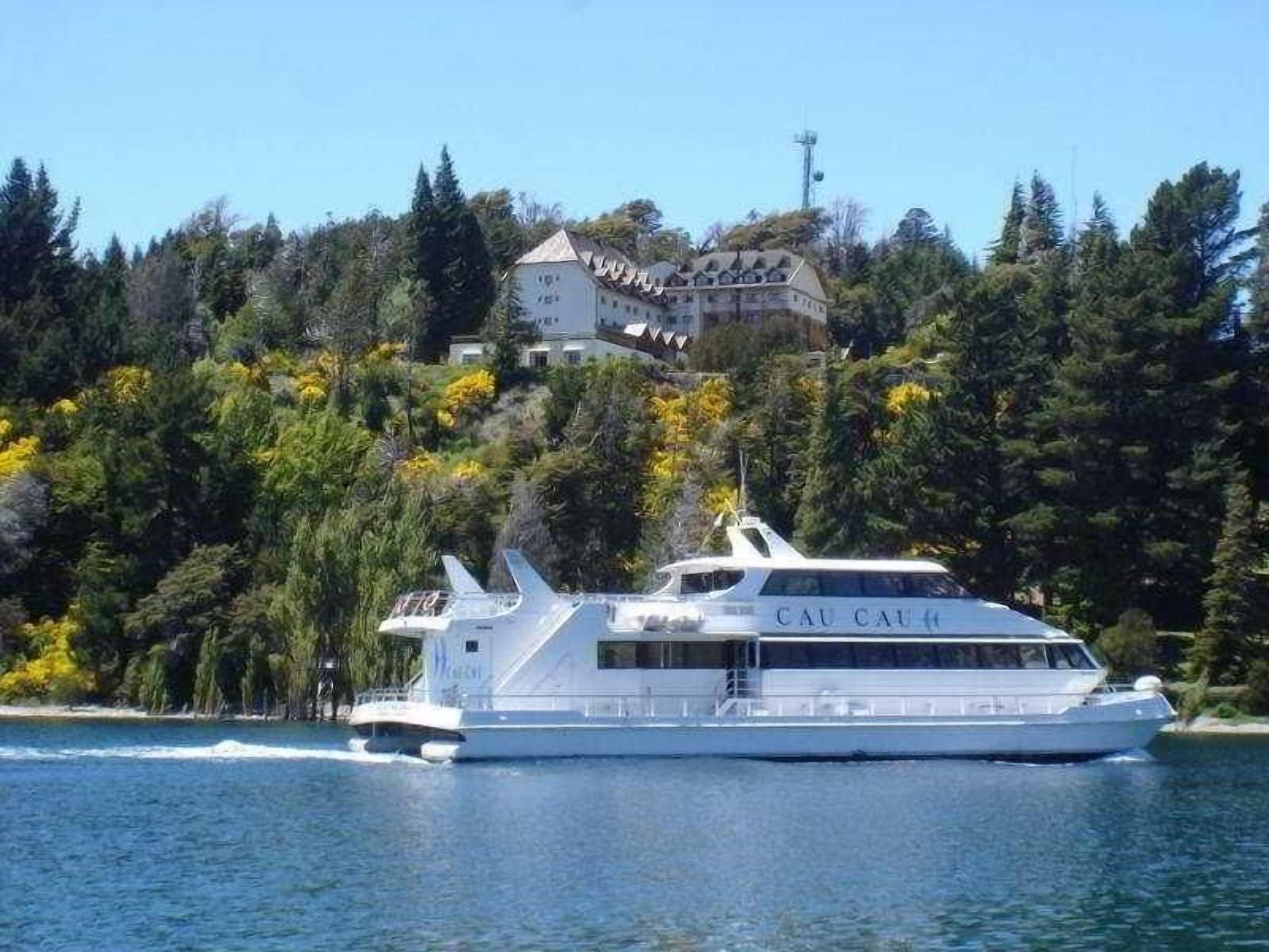 Un gran barco crucero blanco llamado CAU CAU, con distribución de camarotes y vista a una colina boscosa con una casa al fondo.