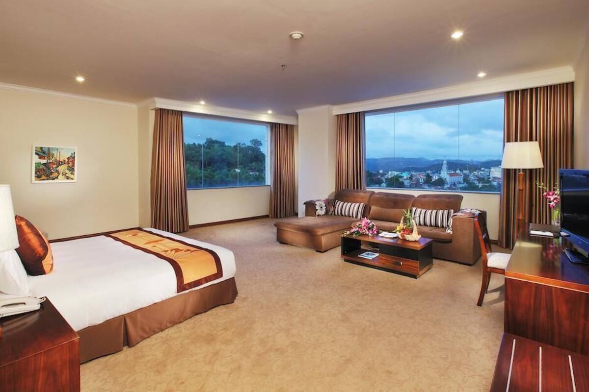 A hotel room with a bed, a sofa, a coffee table, a TV, and large windows offering a city view.