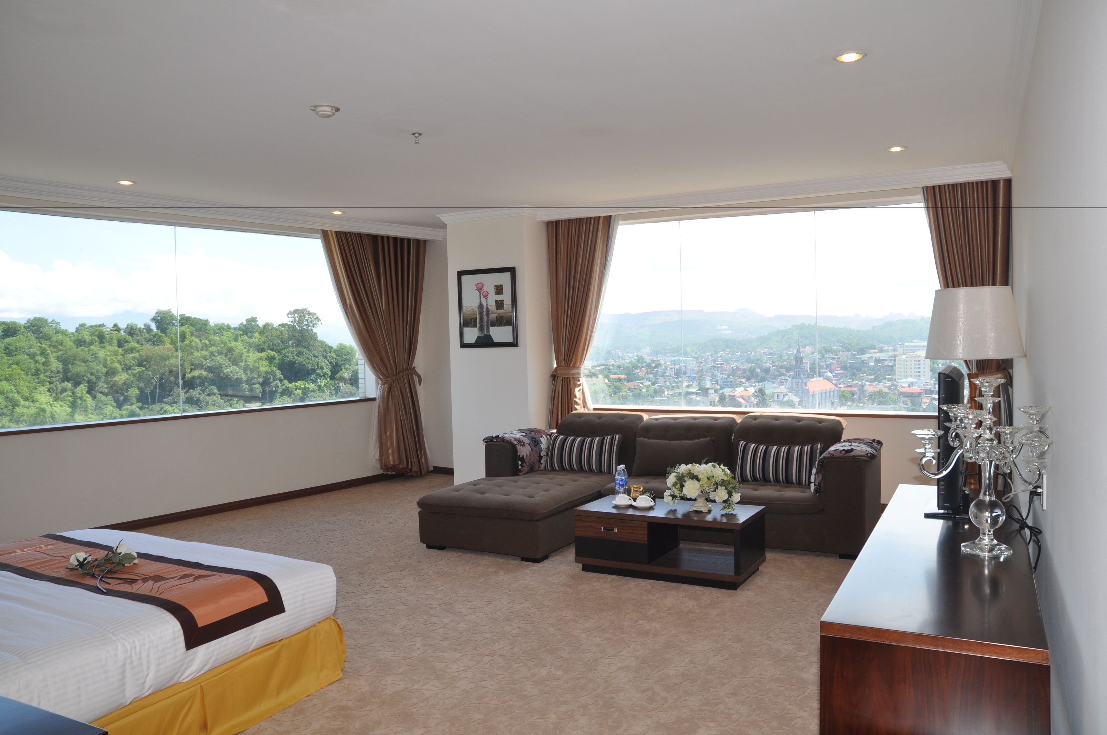 Une chambre d’hôtel avec un grand lit, un canapé, une table basse et une vue sur le paysage urbain grâce à de grandes fenêtres.