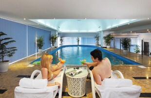 Zona de piscina con dos personas sentadas en una mesa, una piscina y plantas en macetas.