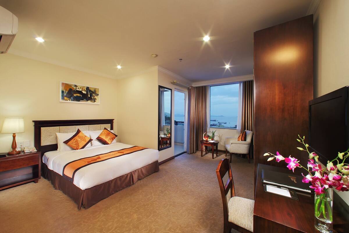 A hotel room with a large bed, a desk, a chair, a TV, and a window with a view.