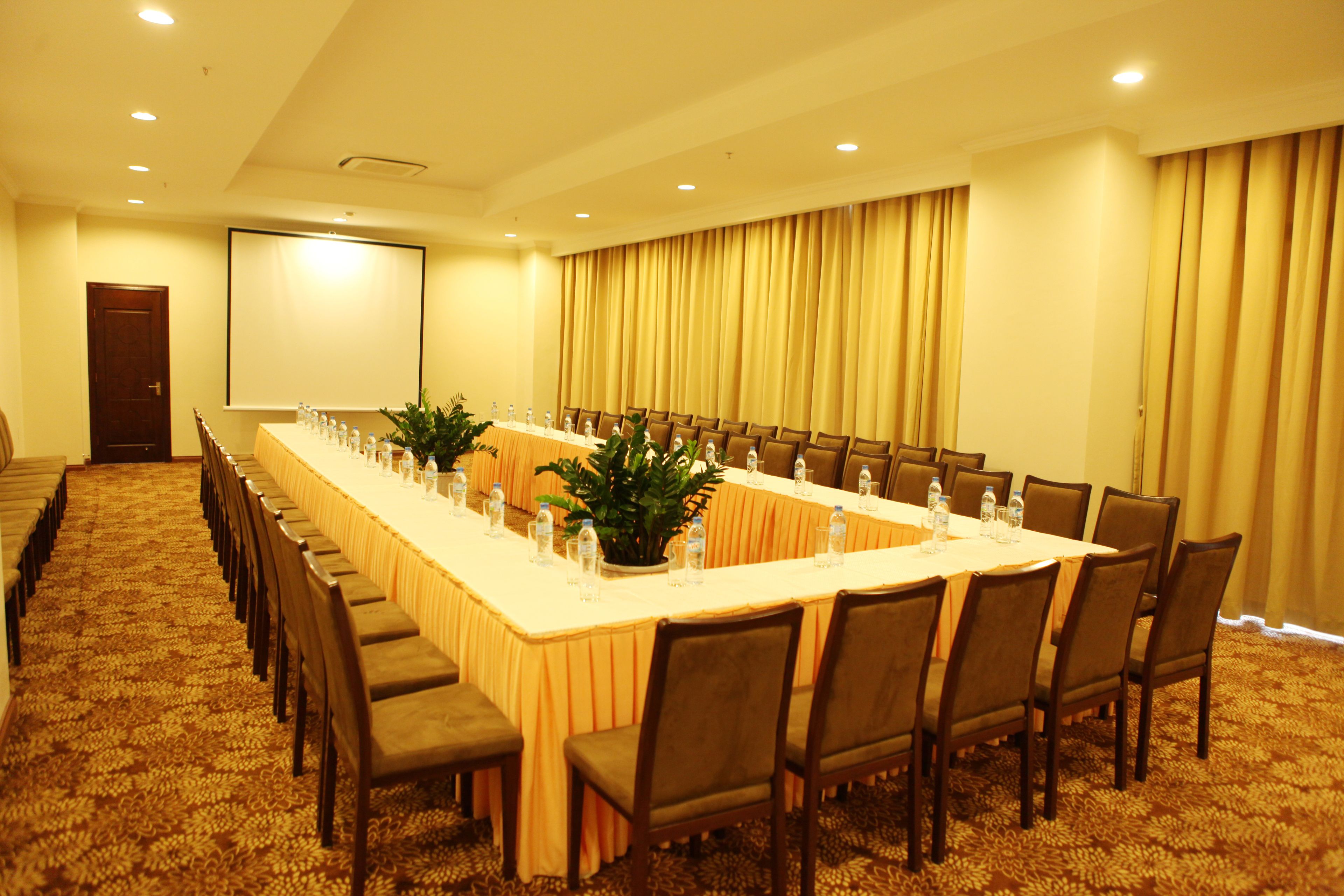 Uma sala de conferências com uma mesa comprida, cadeiras, garrafas de água e uma tela de projetor.