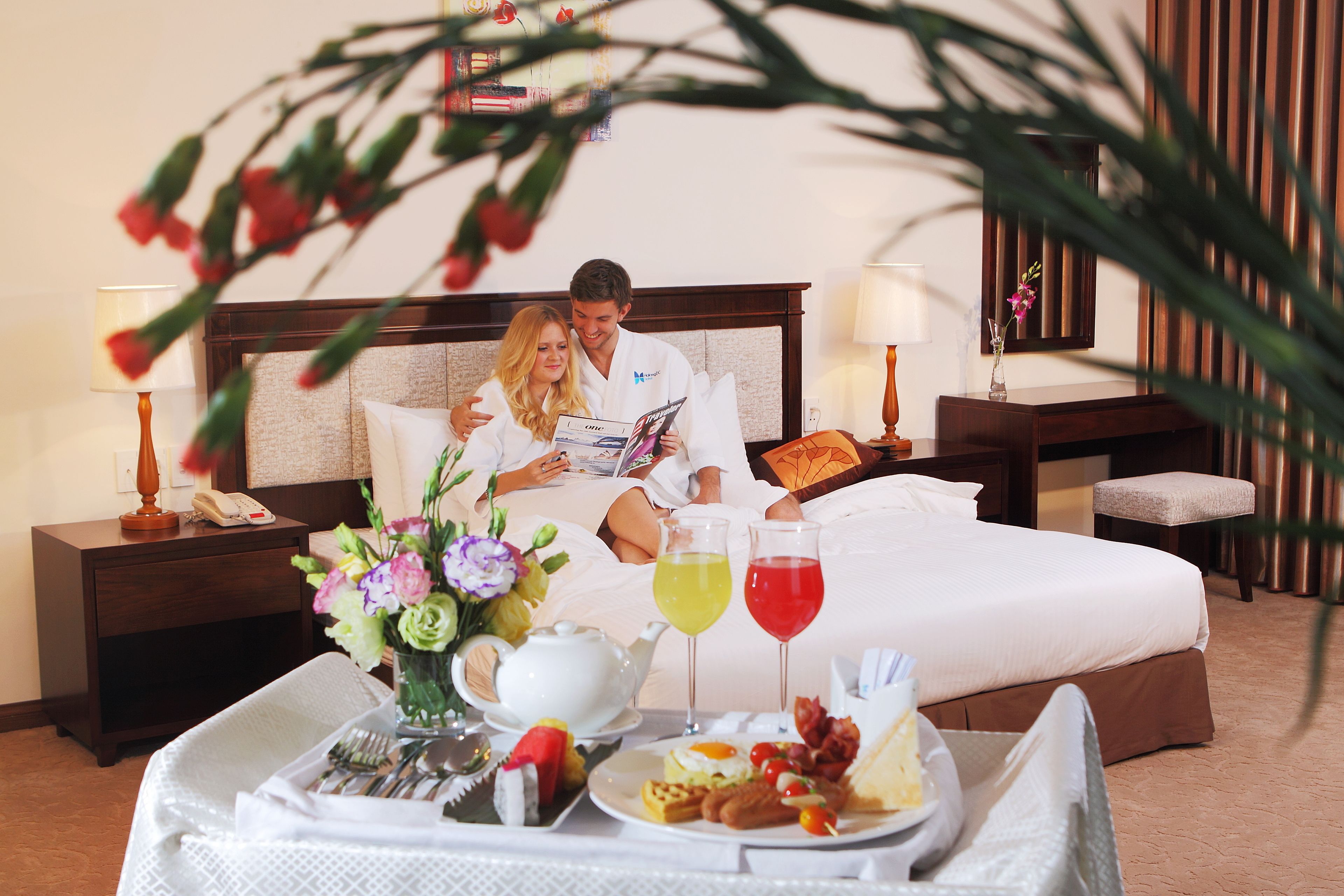 Una habitación de hotel con cama, mesitas de noche, una bandeja con alimentos para el desayuno y un jarrón con flores.