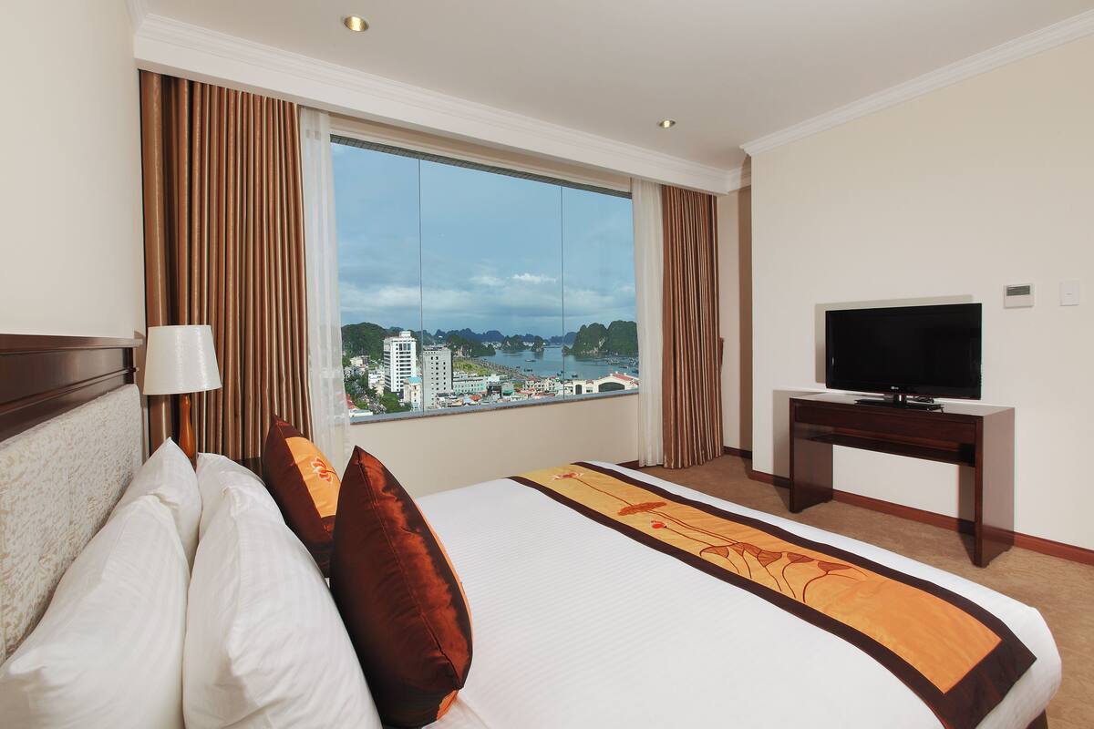 A hotel room with a large window, a bed with white and brown bedding, a television on a wooden stand, and a view of a cityscape.
