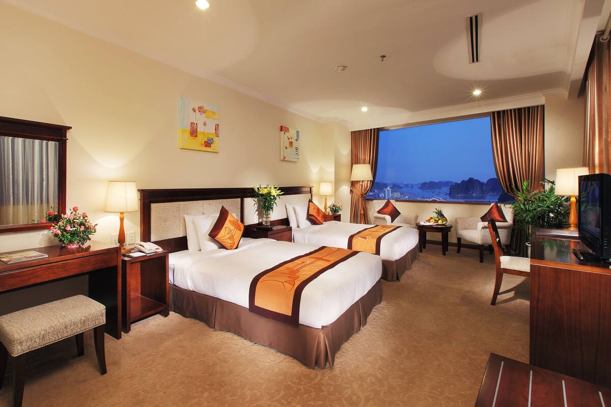 A hotel room with two beds, a desk, a chair, a TV, and a large window with a view.