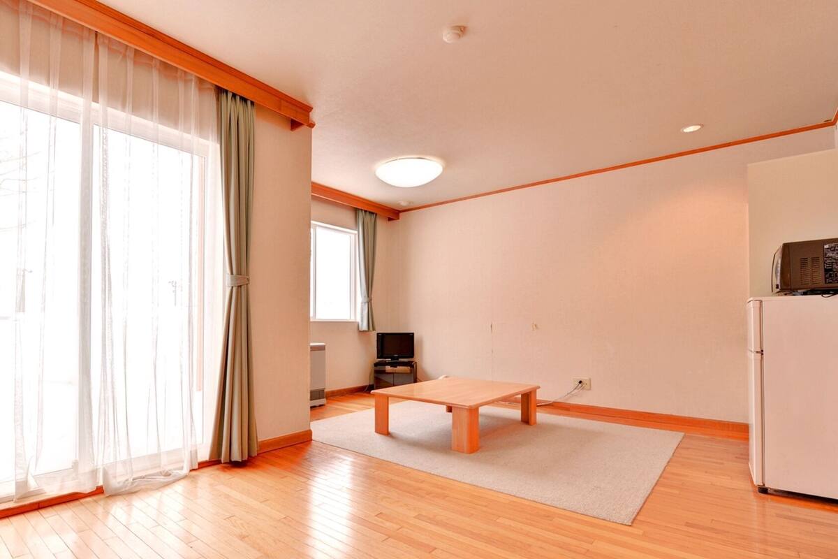 A spacious living room with a wooden floor, a central table, a television, and a refrigerator.