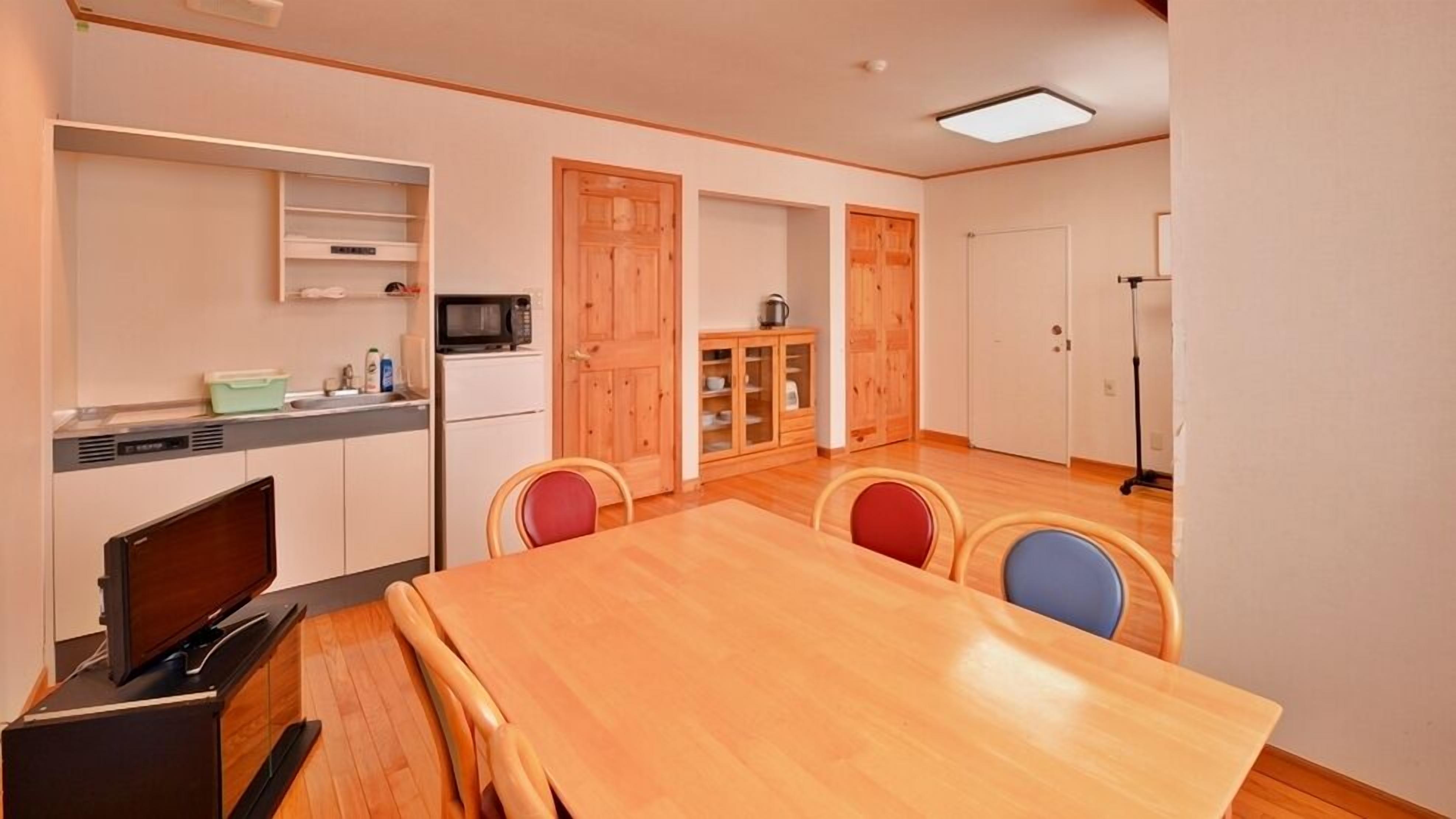 Cocina compacta con microondas, un lavamanos y un mueble pequeño. Hay una mesa de comedor con cuatro sillas y una puerta de madera que da a otra habitación.