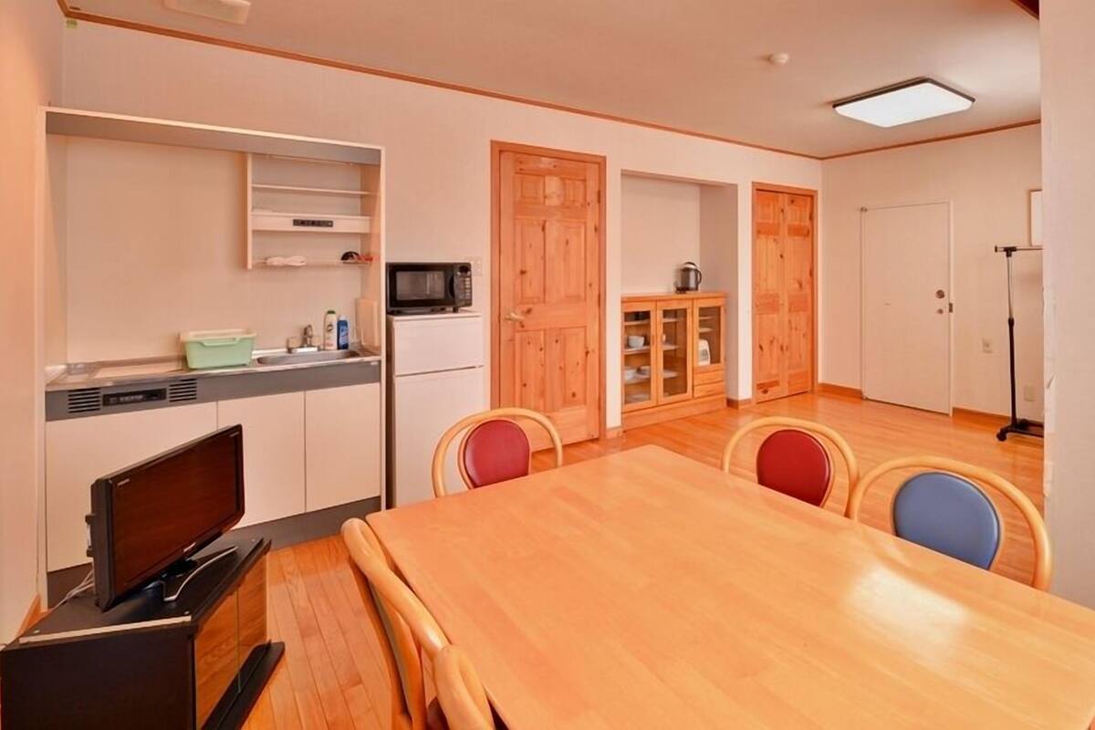 A compact kitchen with a microwave, a sink, and a small cabinet. There is a dining table with four chairs, and a wooden door leading to another room.