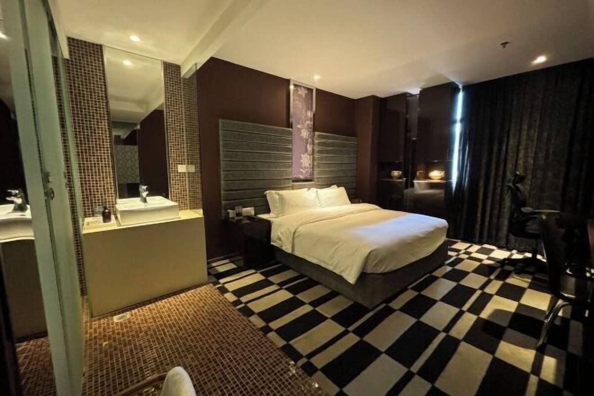 A hotel room with a checkered floor, a bed with white linens, a large mirror, and a dark-colored headboard.