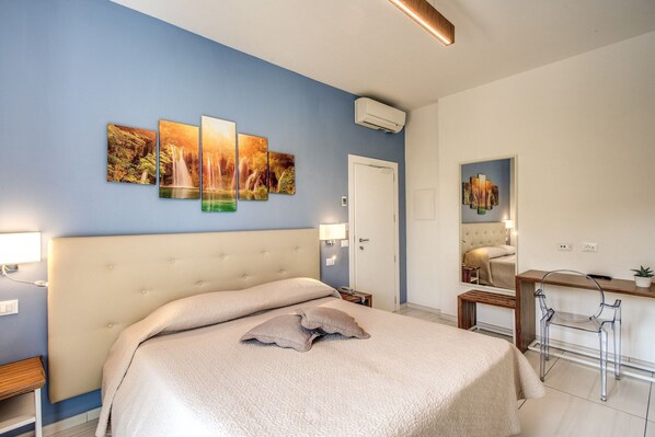 A bedroom with a large bed, a desk, and a chair. There is a door and an air conditioning unit on the wall.