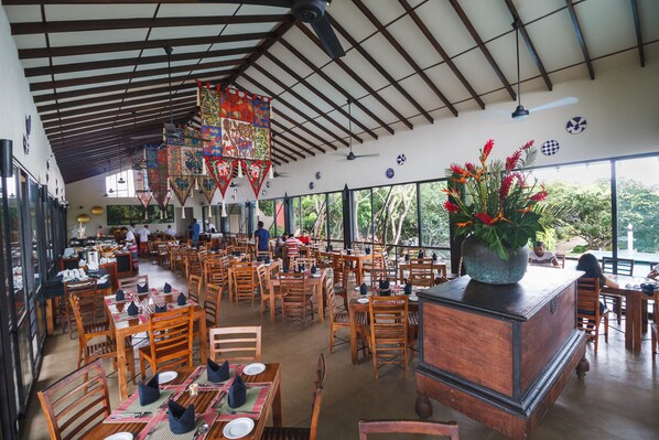 Un restaurant spacieux avec des tables et des chaises en bois, un plafond haut avec des poutres apparentes et de grandes fenêtres offrant une vue sur l’extérieur.