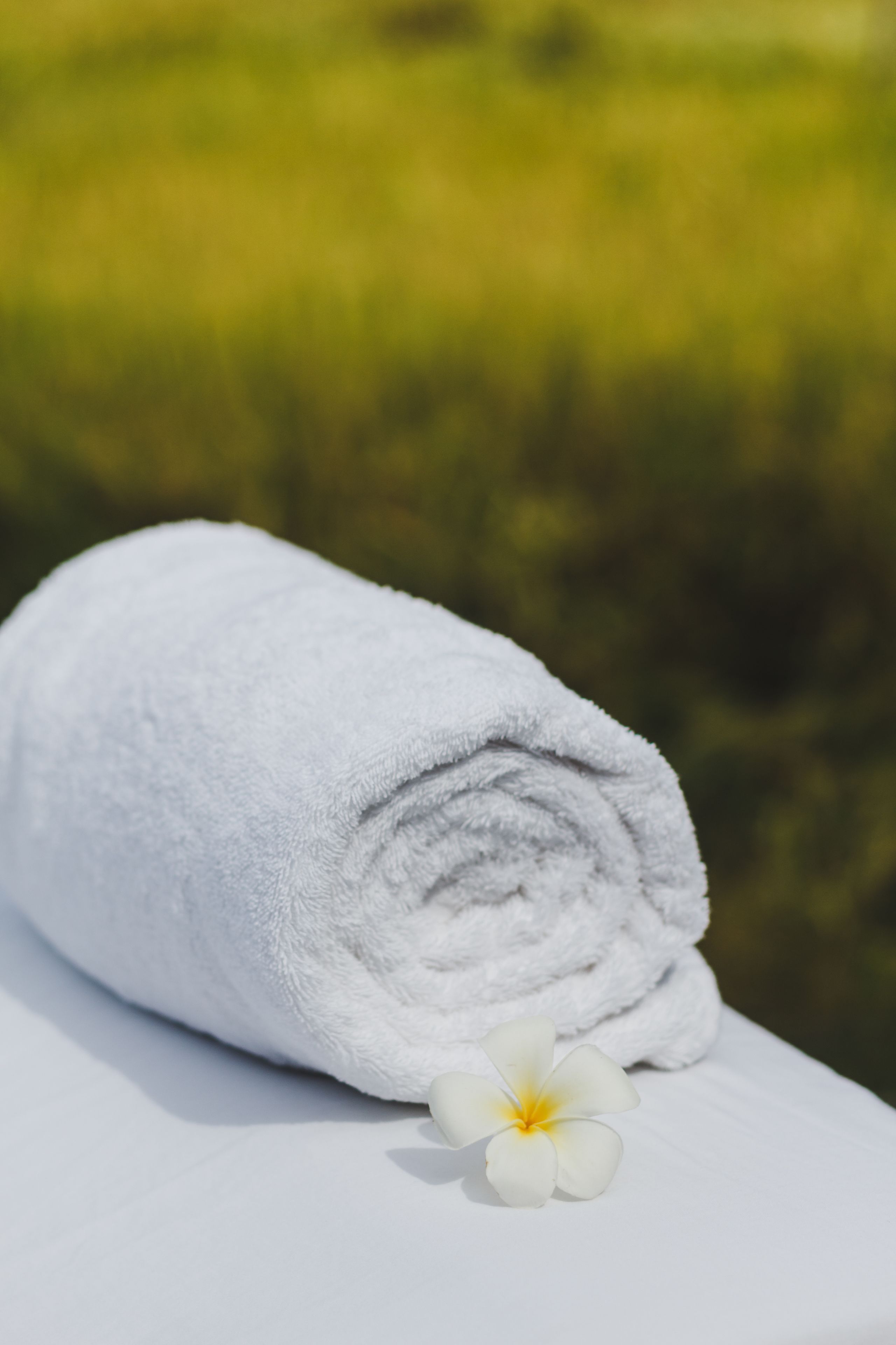 A rolled white towel and a white flower on a white surface with a blurred green background.