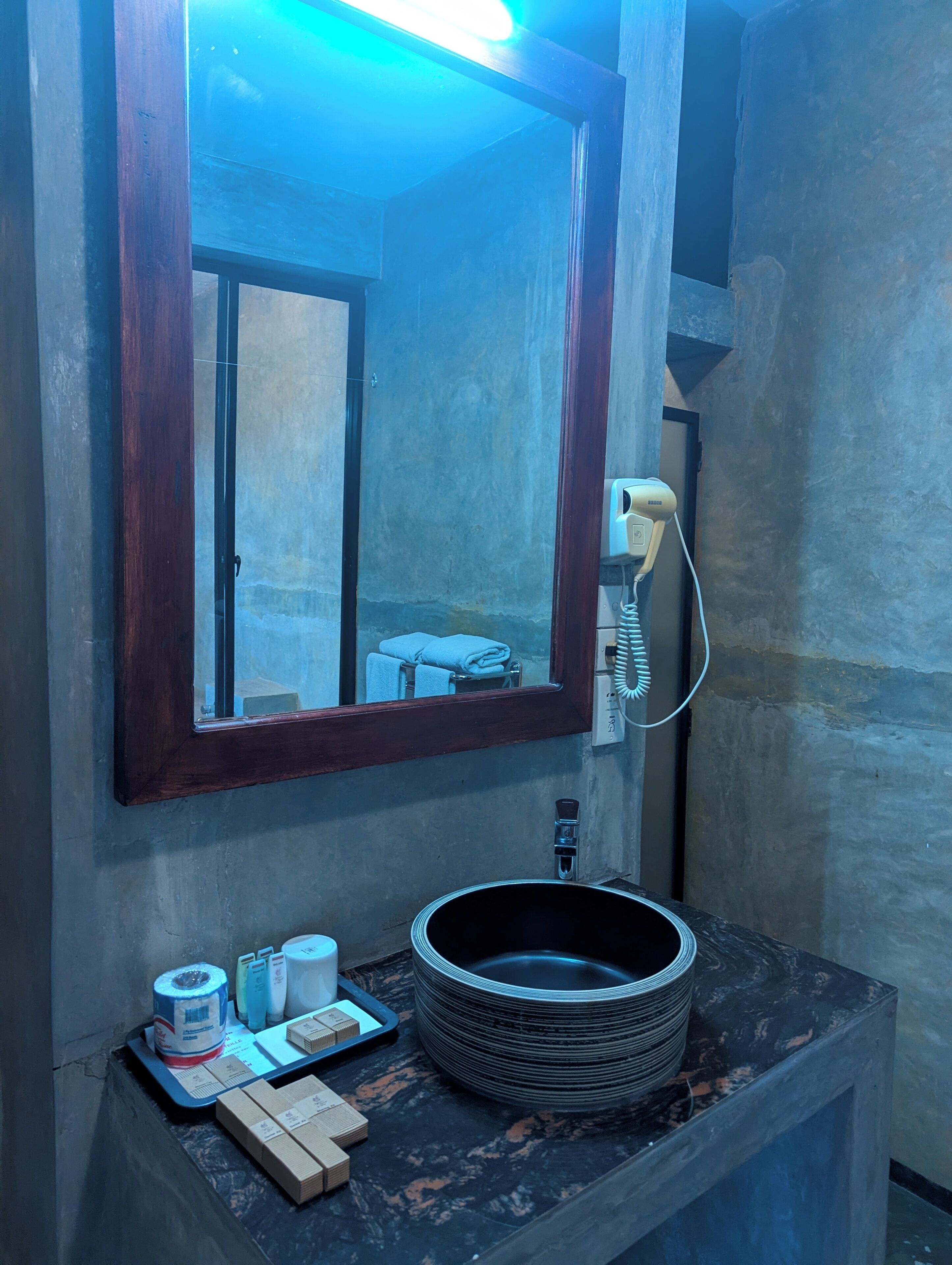Un baño con espejo de marco de madera, un lavabo con lavamanos negro y un perchero para toallas.