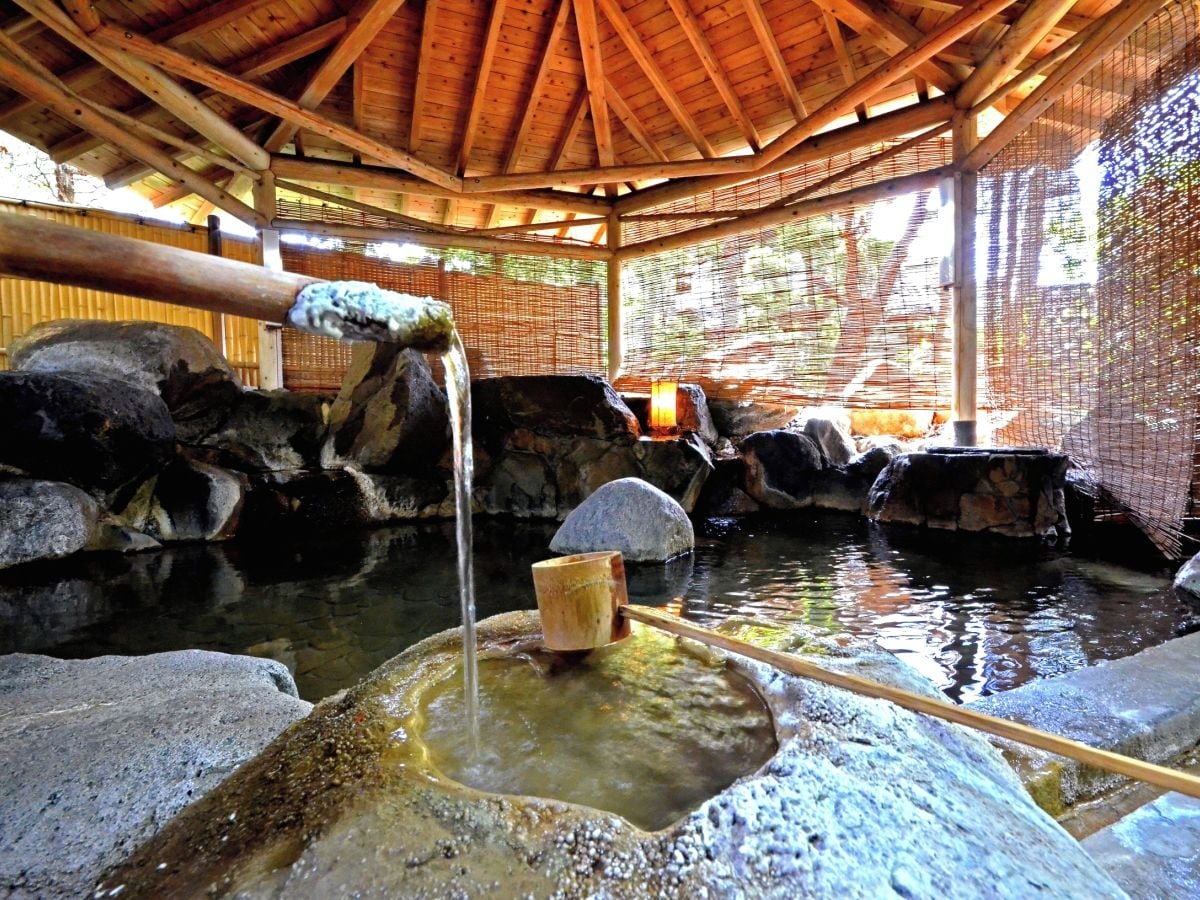 Un bain traditionnel dans une source d’eau chaude, avec un toit en bois, des murs en pierre et un seau en bois.