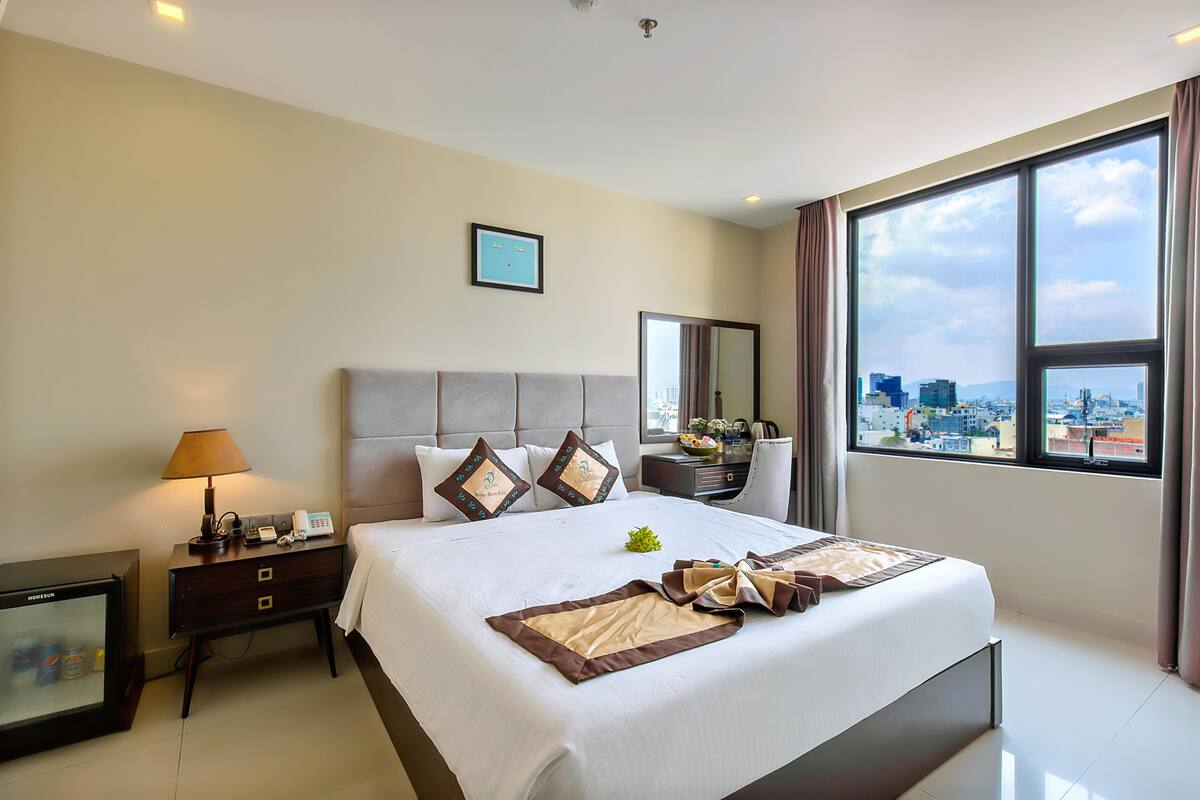 Habitación de hotel con una cama grande, una mesita de noche con lámpara, televisión y vistas a la ciudad desde la ventana.