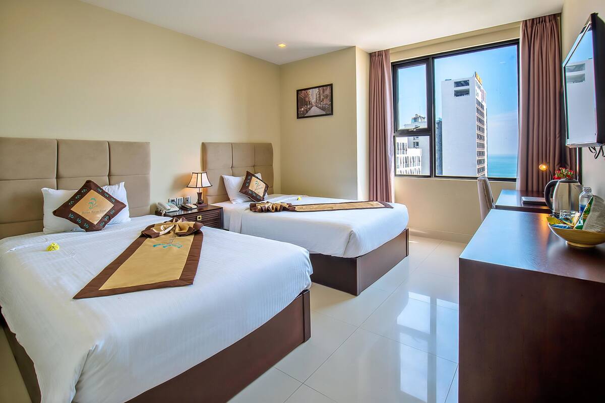 Habitación de hotel con dos camas, un escritorio, un televisor y un amplio ventanal con vistas a la ciudad.