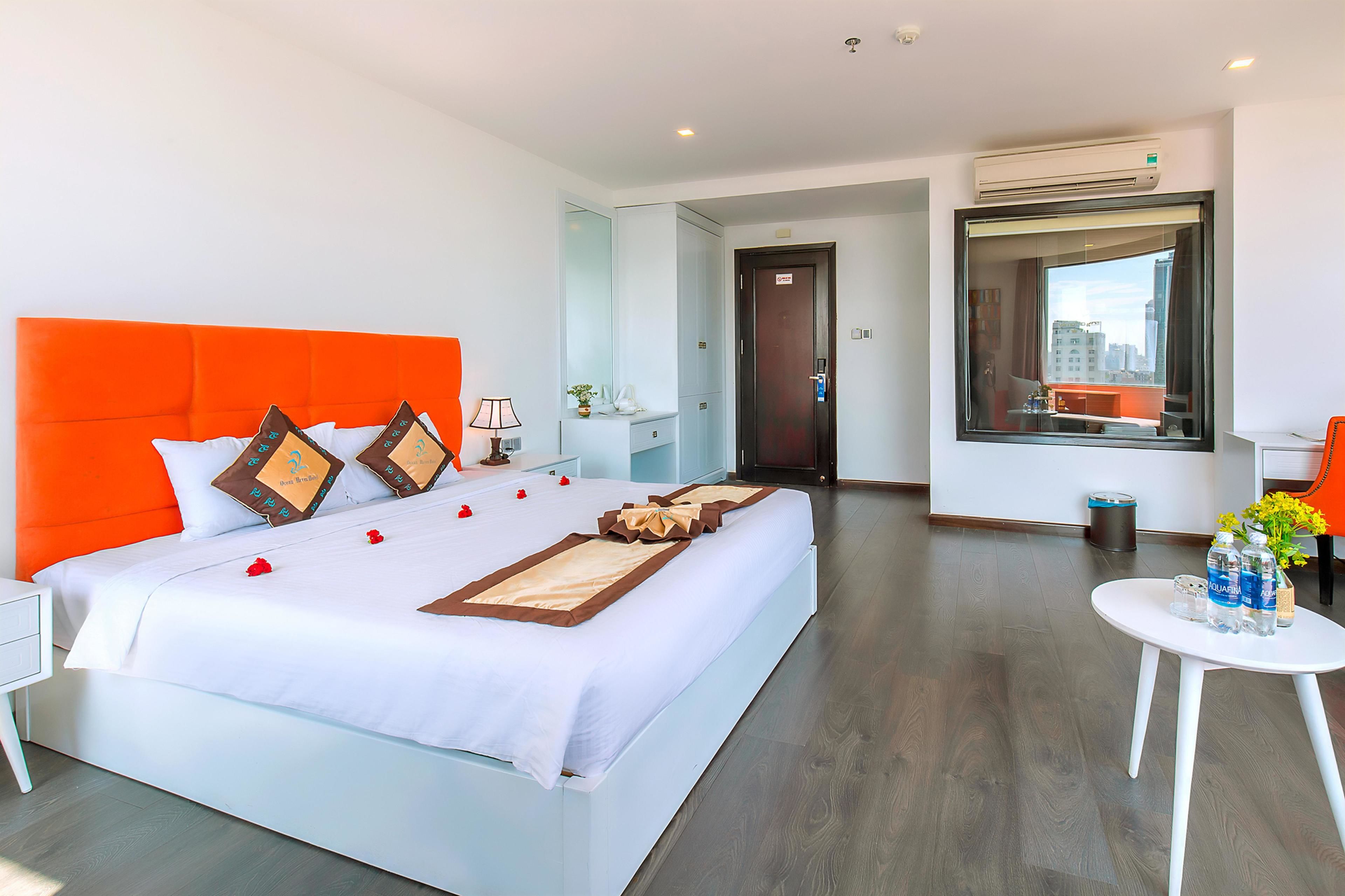 A modern hotel room with a large bed, white bedding, orange headboard, and a view of the city through a large window.
