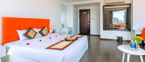A modern hotel room with a large bed, white bedding, orange headboard, and a view of the city through a large window.