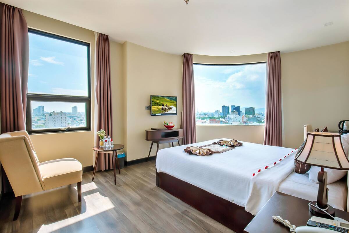 Habitación de hotel con una cama grande, un escritorio, una silla, un televisor y vistas a la ciudad.
