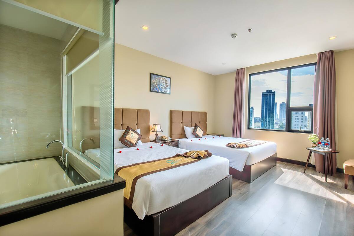 Habitación de hotel con dos camas, un ventanal con vistas a la ciudad, un baño con cabina de ducha de vidrio y una pequeña mesa con una planta y botellas de agua.