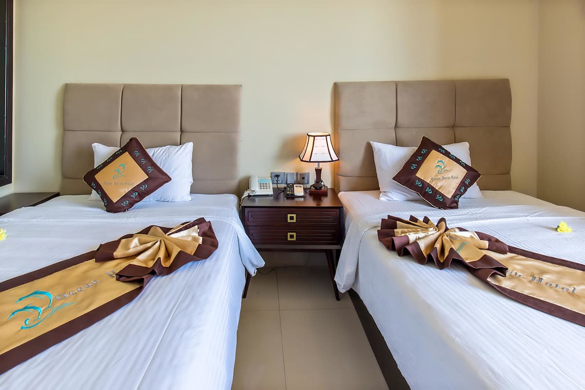 Habitación de hotel con dos camas, cada una con cabecero marrón, ropa de cama blanca y almohadas decorativas. Entre las camas, una mesita de noche con lámpara y teléfono.