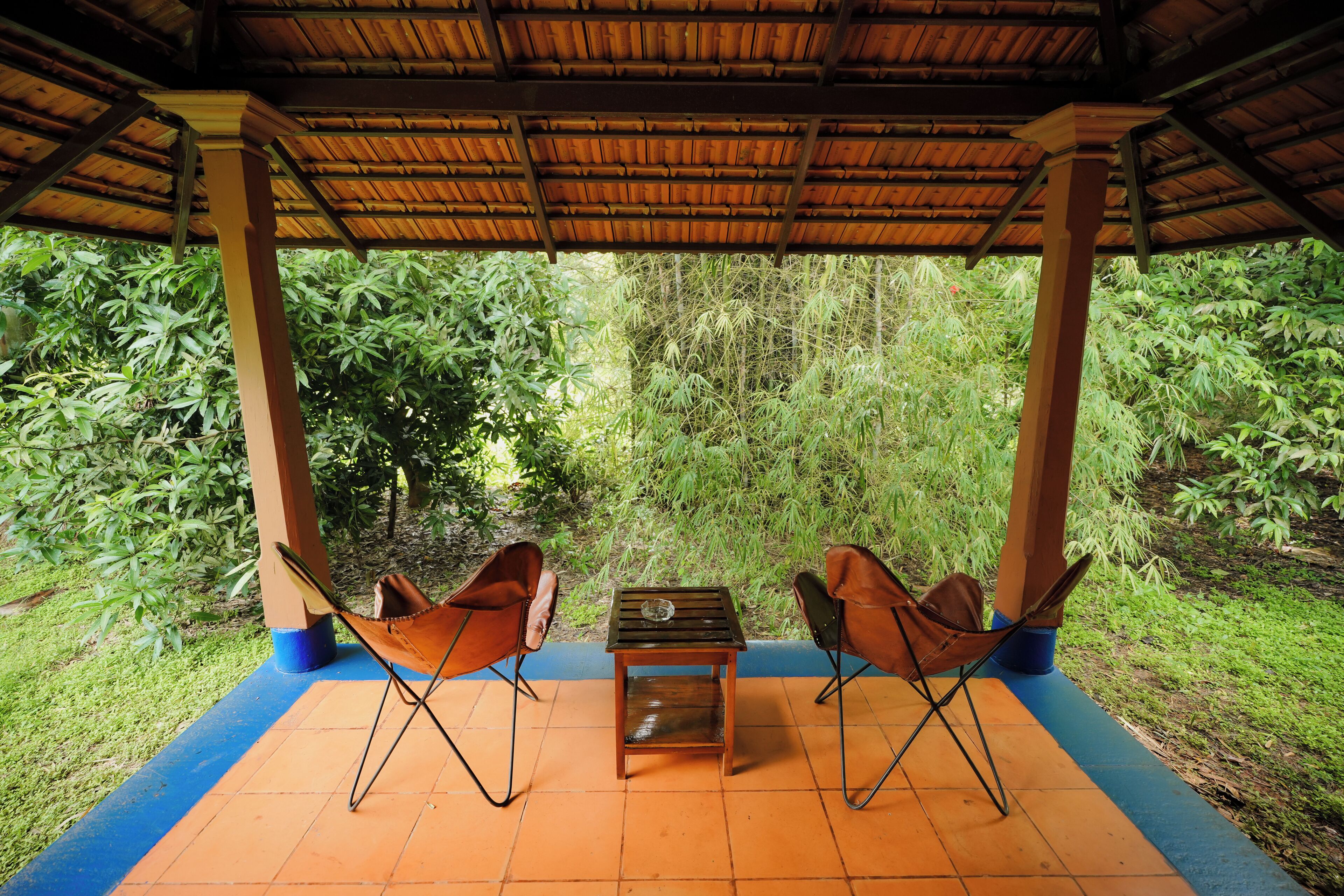 A covered outdoor seating area with two chairs and a small table, surrounded by lush greenery.