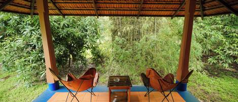 A covered outdoor seating area with two chairs and a small table, surrounded by lush greenery.