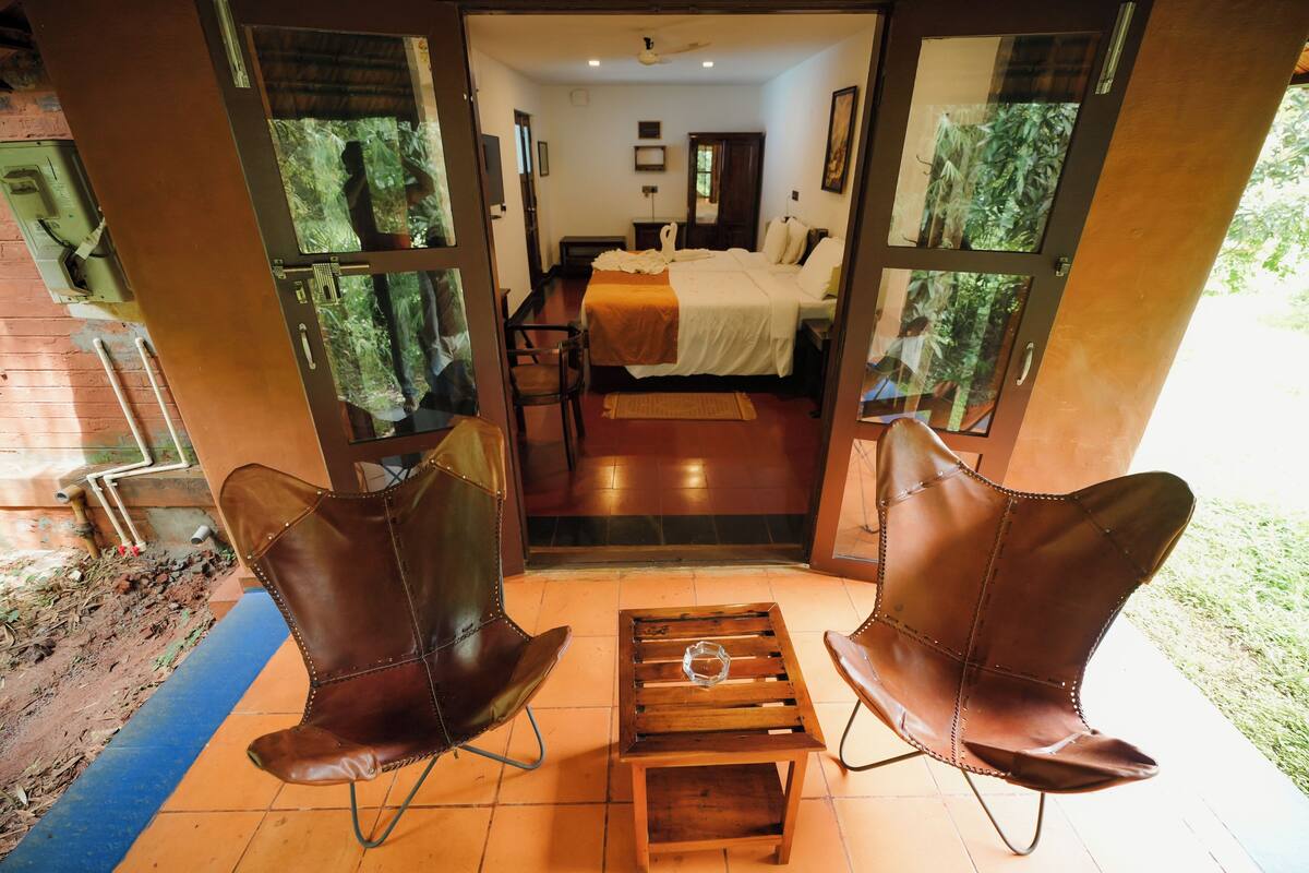 A room with a bed, two leather chairs, a small wooden table, and a view through a glass door.