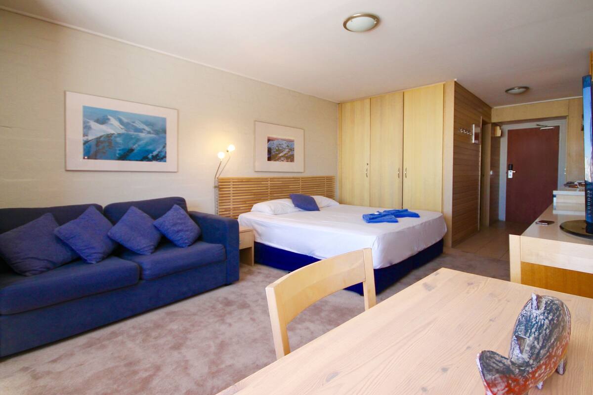 A room with a bed, a wooden wardrobe, a blue sofa, a wooden table, and two framed pictures on the wall.