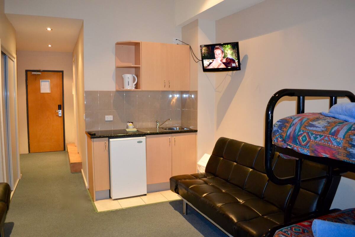A compact room with a kitchenette, a sofa, and a television mounted on the wall.