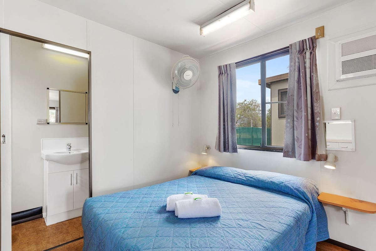 Un dormitorio con cama, mesita de noche, ventana con cortinas, ventilador en la pared y baño con lavamanos y espejo.
