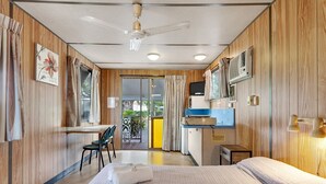 A compact hotel room with a bed, a desk, and a chair. There is a small kitchenette with a microwave and a sink. The room has wooden paneling and a ceiling fan.