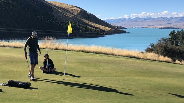 Un campo de golf con una bandera, una persona de pie y otra sentada sobre el césped.