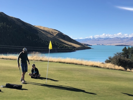 A golf course with a flag, a person standing, and another person sitting on the grass.